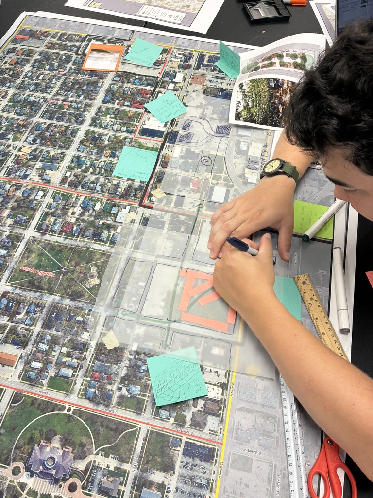 A workshop participant sketches neighborhood ideas over an aerial map, surrounded by notes and courtyard plans.